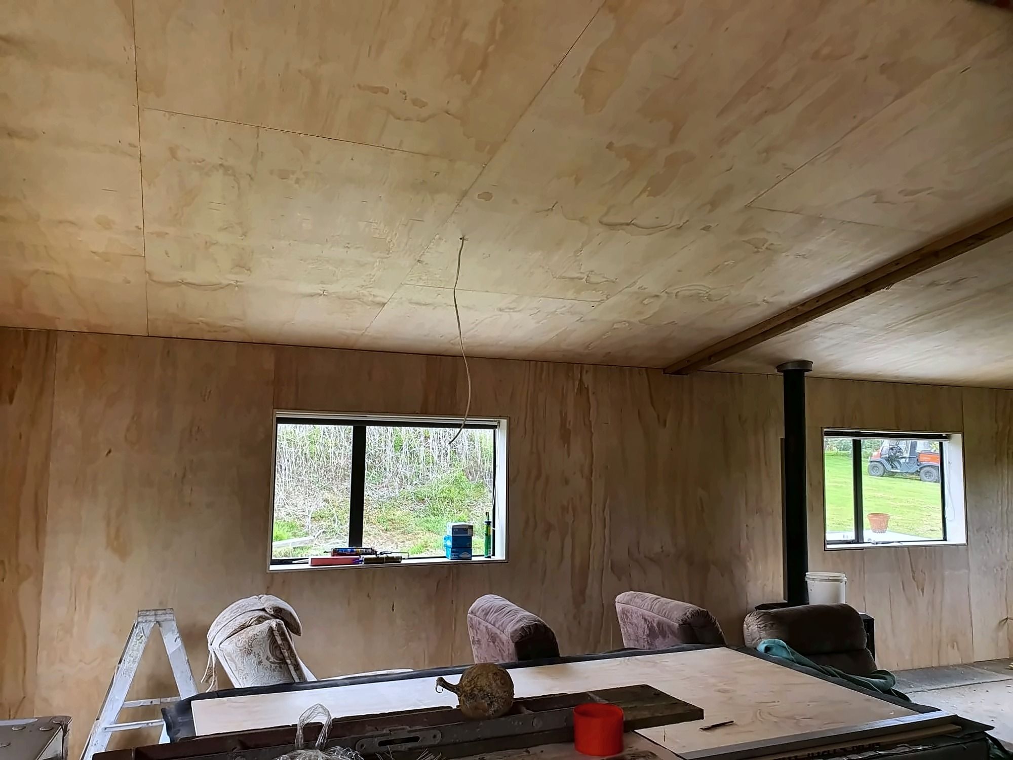 The same room lined in plywood on the walls and ceiling, with two windows and a wood burner flue.