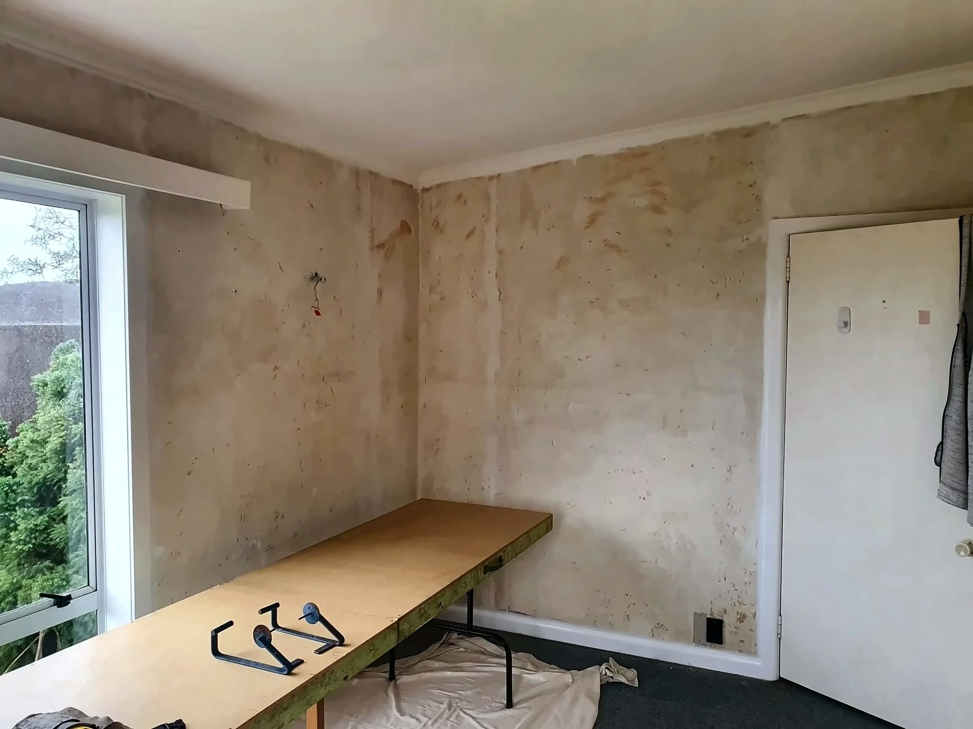 Bedroom walls stripped back to a bare, patchy surface, with a work table set up in the middle of the room.